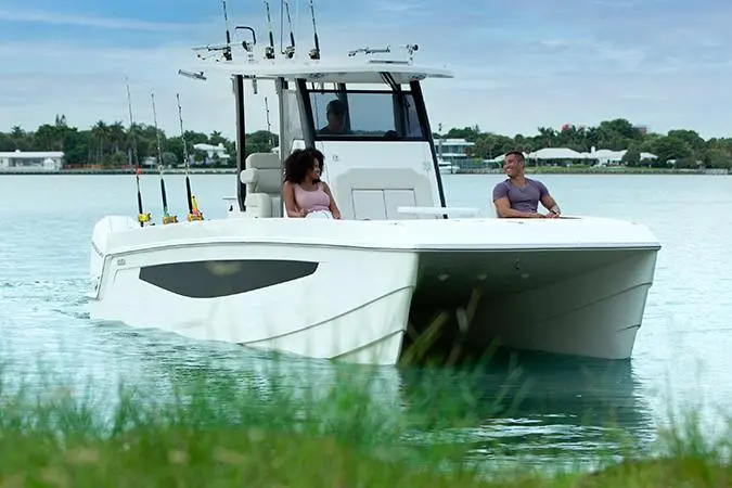 A white fishing boat with two people seated inside, surrounded by calm water and greenery, under a cloudy sky.
